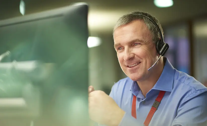 a call centre phone operative in his mid 40s chats on the phone at his desk . He is explaining something to the person on the phone in a friendly manner . behind him a defocussed office interior can be seen . This could be a call center or an office worker chatting to a customer.