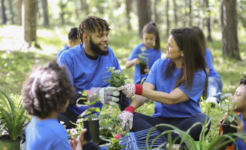 A group of environmental volunteers in matching bright blue t-shirts plant flowers, trees, and plants at a local park during the spring.