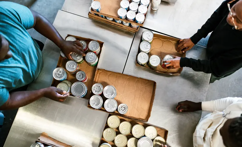 A group of young adult friends volunteer time working at a food bank, processing donations of packaged food products and clothing. Overhead view of them sorting canned food.