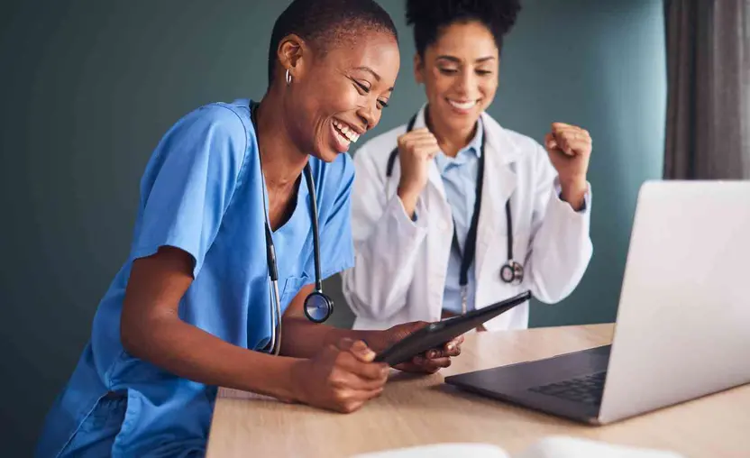 Two female physicians are gathered around a laptop while smiling and celebrating.