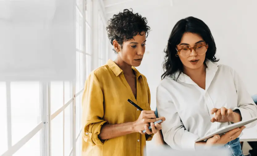 Two women stand side-by-side and take notes on a pad and tablet as they discuss their organization’s impact measurement.