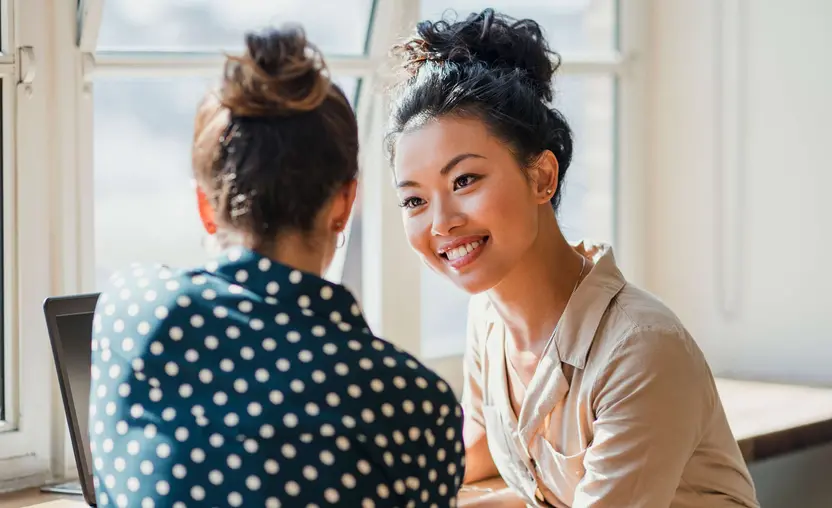 A woman smiles at a coworker in front of a window and laptop.