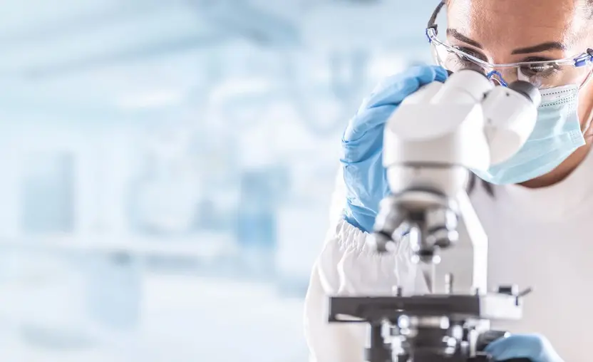 A female scientist is wearing a lab coat, goggles, gloves, and a mask while looking into a microscope. 