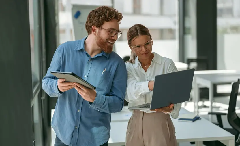 A male and a female employee are standing in an office. The female employee is holding a laptop, and the male employee is holding a tablet. They are both looking at the laptop screen and smiling. 