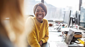 A nonprofit professional sits at a desk and smiles at her colleague as they discuss the importance of sustainers to their organization.