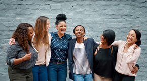 A diverse group of women standing in front of a brick wall with their arms around each other. 