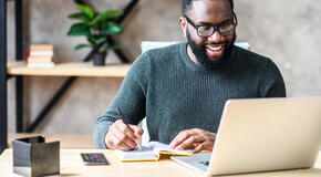 Cheerful African-American male student or worker in glasses is watching online lectures or webinars and writing notes in a notebook