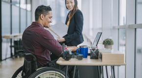 Man in wheelchair working on a computer while talking with a female colleague.