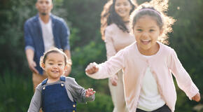 Two young children with brown hair are smiling and running, and their parents are in the background behind them.