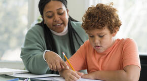 A mother helps her child do their homework at their kitchen table.