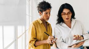 Two business women having a discussion, they're standing in an office and using a tablet. Professional women making a to do list as part of their project planning.