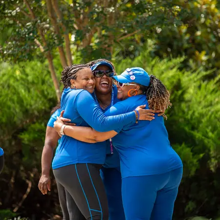 Three Black women wearing their royal blue GirlTrek member shirts embrace and laugh together. They are out on a GirlTrek walk together, it’s a sunny day, and there are trees in the background.