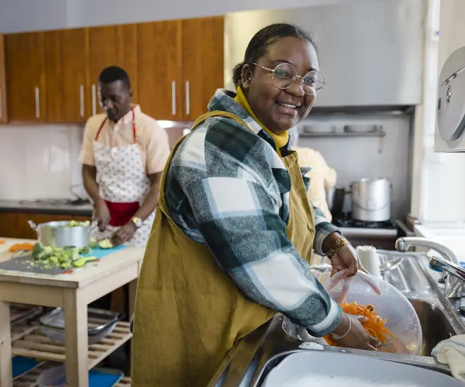 A shot of volunteers cleaning and washing up in the kitchen at a warm hub/food bank which is a safe place for people to enjoy a warm and friendly environment in the community with the current cost of living increasing. They have made hot dinners for the visitors, one young woman is smiling and looking at the camera.