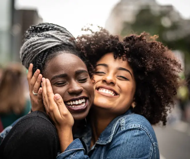 Two African American women stand outdoors smiling and hugging