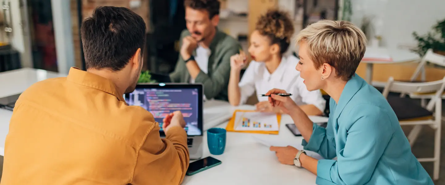 A team of four people gathers around a table with a laptop and papers to start a nonprofit organization website.