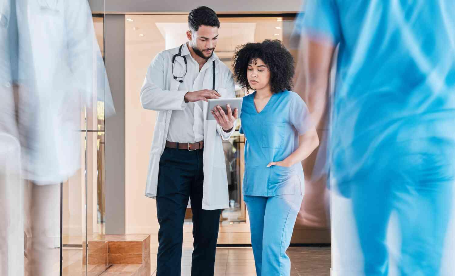 A male and a female physician are standing in the hallway of a hospital reviewing medical records on a tablet. 