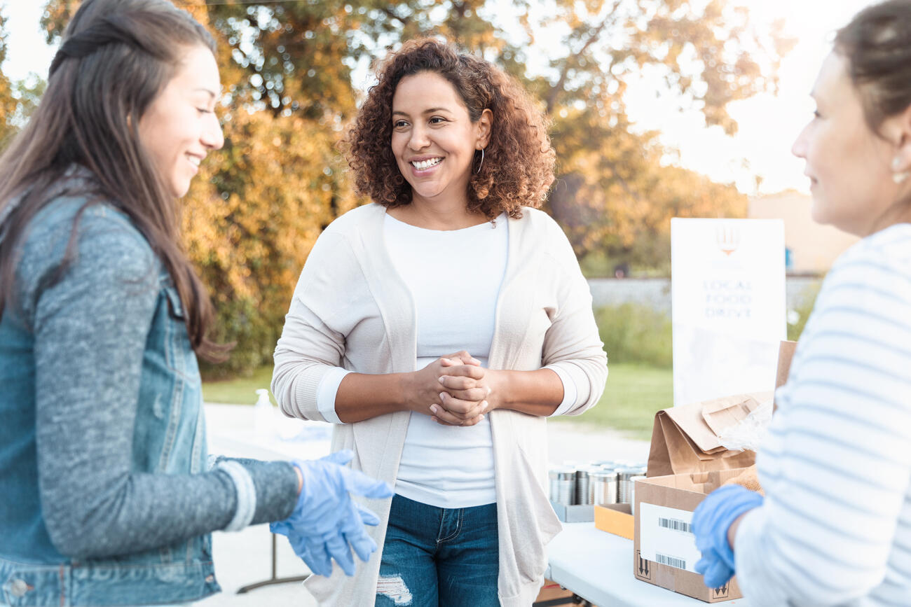 Three female friends enjoy volunteering together during a community food drive. They are collecting food donations for people impacted by the coronavirus pandemic.