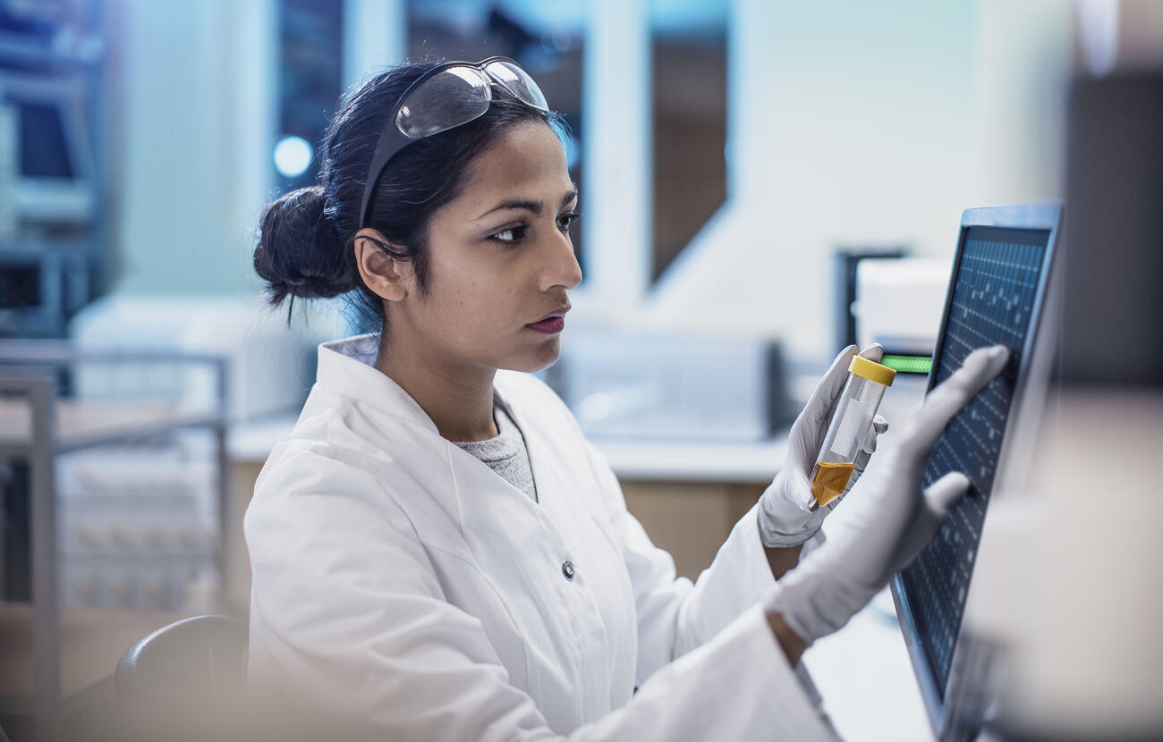 A scientist works in a lab using a touch screen computer. She has long dark hair and is wearing a white coat.