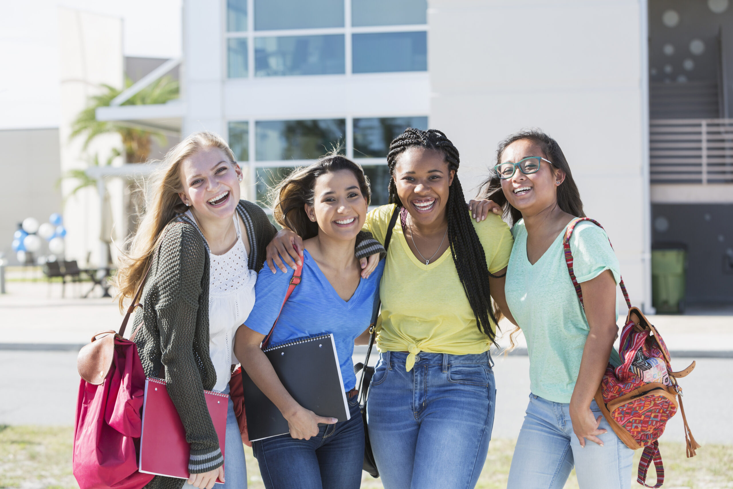 A group of four multi-ethnic teenage girls, 18 years old, standing in front of a school building carrying book bags and notebooks. They are high school seniors or freshmen in college.