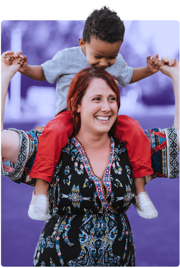 A woman carries her toddler son on her shoulders and smiles while enjoying a relaxing walk in a park.