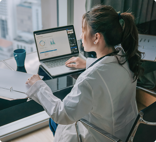 A doctor works at her desk on a laptop while reviewing paperwork.