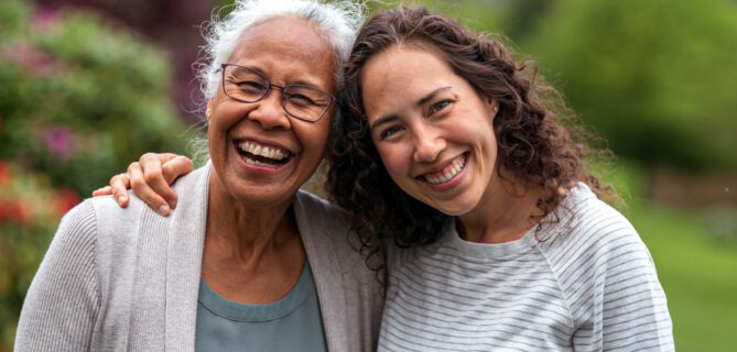 An elderly mother and her grown daughter embrace and smile at the camera