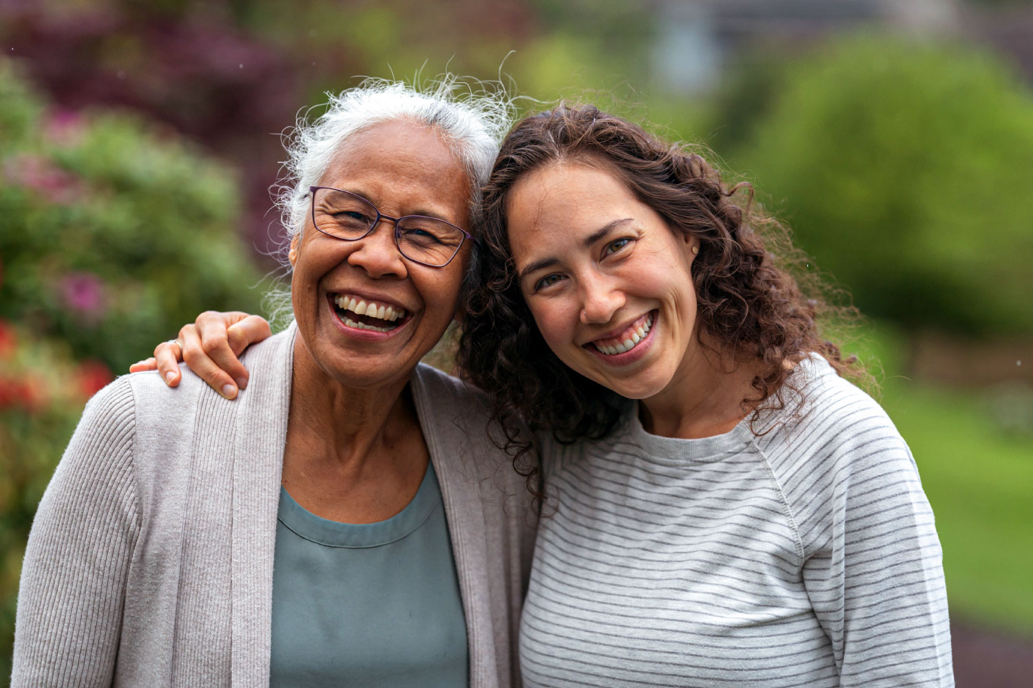 An elderly mother and her grown daughter embrace and smile at the camera